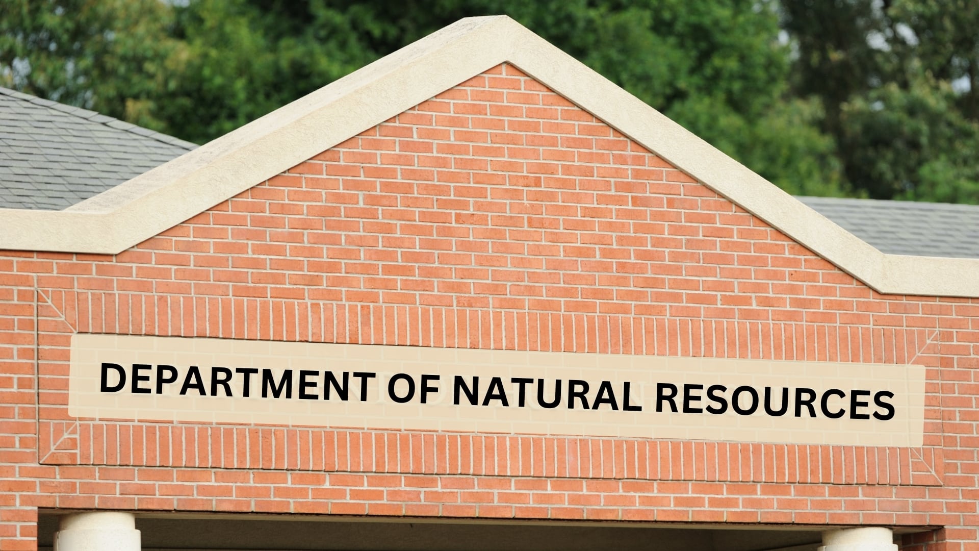 The image shows the front facade of a brick building with a triangular roof. On the front of the building, there is a large sign that reads "DEPARTMENT OF NATURAL RESOURCES" in bold, black letters. The background features greenery, indicating that the building might be located in a natural or suburban setting.